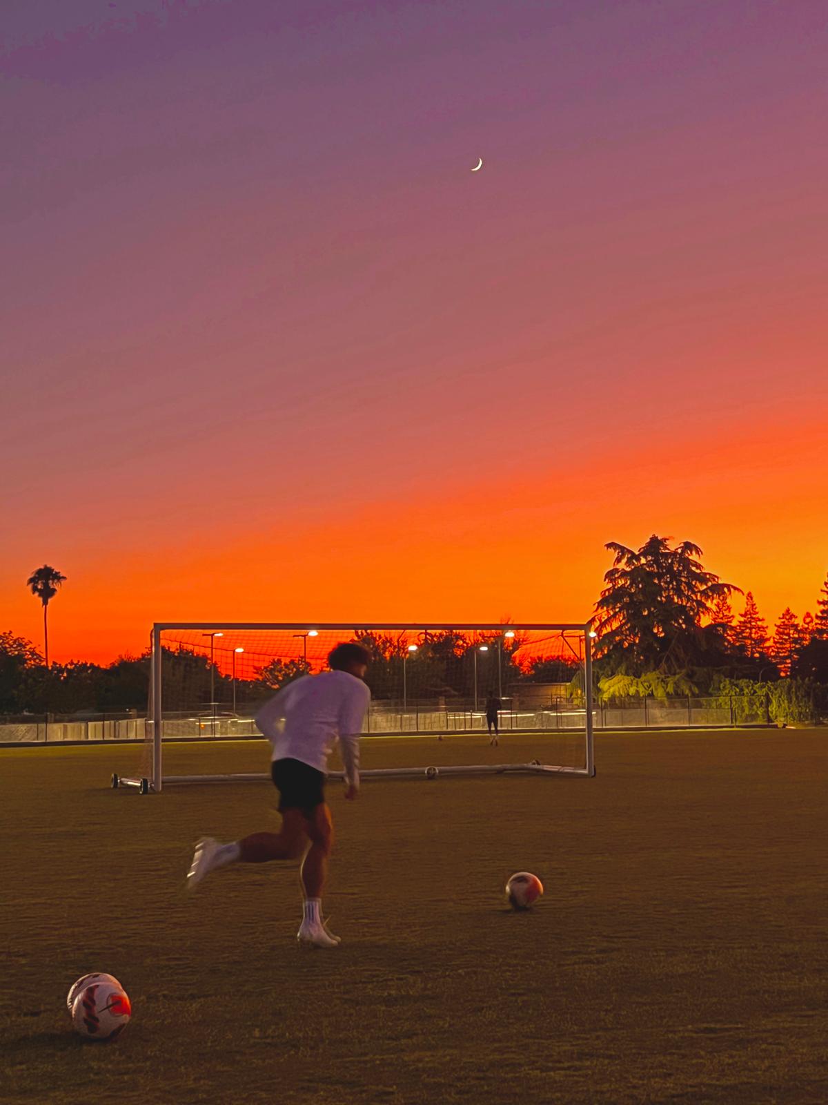 A player running at a ball on a field under an orange sunset.